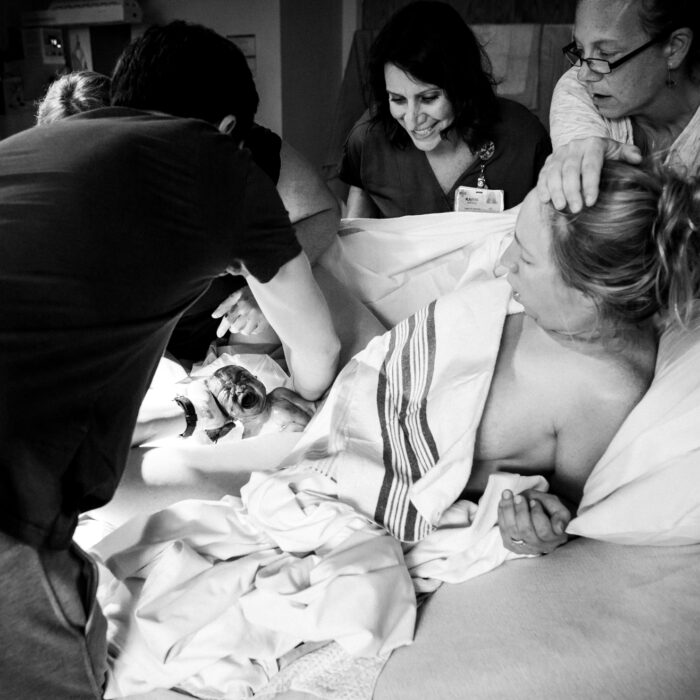 Birthing person in hospital bed, elevated onto elbows. Baby emerging between the birthing person's legs, parent with their back to the viewer is leaning forward catching their baby. Nurse is leaning forward toward birthing person. Doula is holding the neck and top of the birthing person's head.